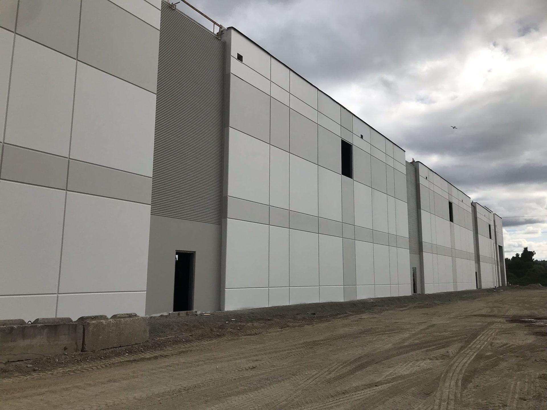 Exterior of a large industrial building under construction, featuring white and gray panels, cloudy sky.