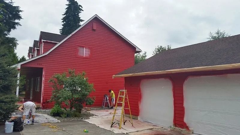 House and garage being painted red, with white sections. Two people use ladders. Exterior setting.