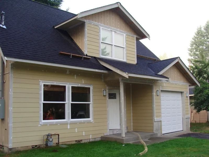 Tan house with dark roof and white trim. Includes a garage, windows, and a small porch.