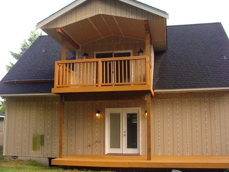 Back of a house with a wooden deck and balcony. Light brown siding, dark roof, French doors.