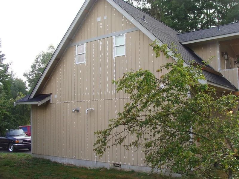 Tan house with vertical siding and two small windows, with patches of white. A truck sits nearby.