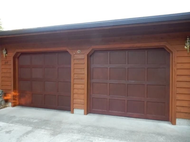 Two dark brown garage doors on a light brown building with a concrete driveway.