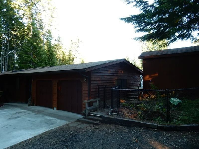 Brown garage with two bays, set in a wooded area, with a concrete driveway and small fence.