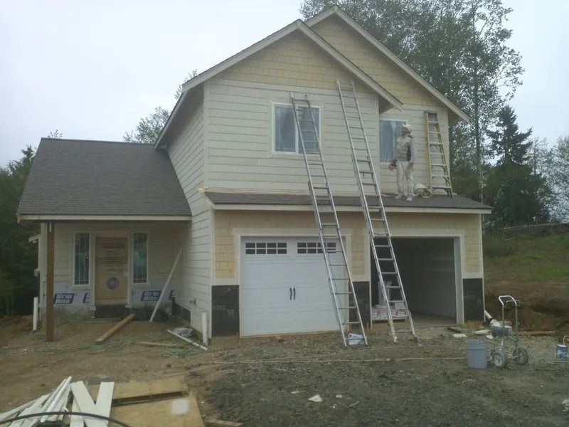 Two-story house under construction, beige siding, two ladders, garage door, person painting, overcast day.