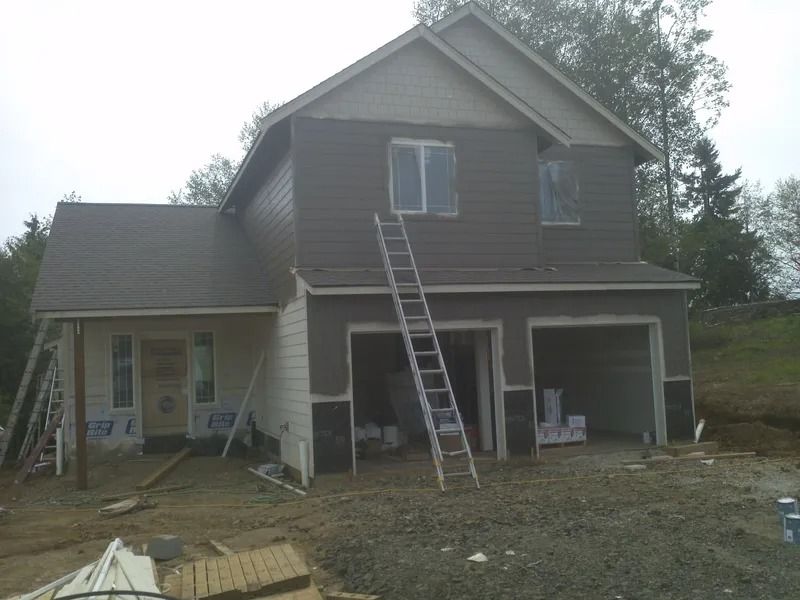 Two-story house under construction with brown siding, two open garage doors, and a ladder leaning against the second story.