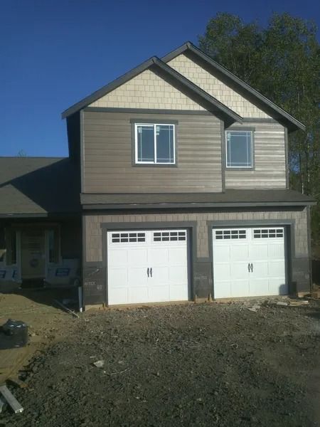 Two-story house with two-car garage. Brown siding, white garage doors, windows, gravel driveway.