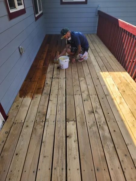 Man applying stain to wooden deck boards, with unfinished section visible.