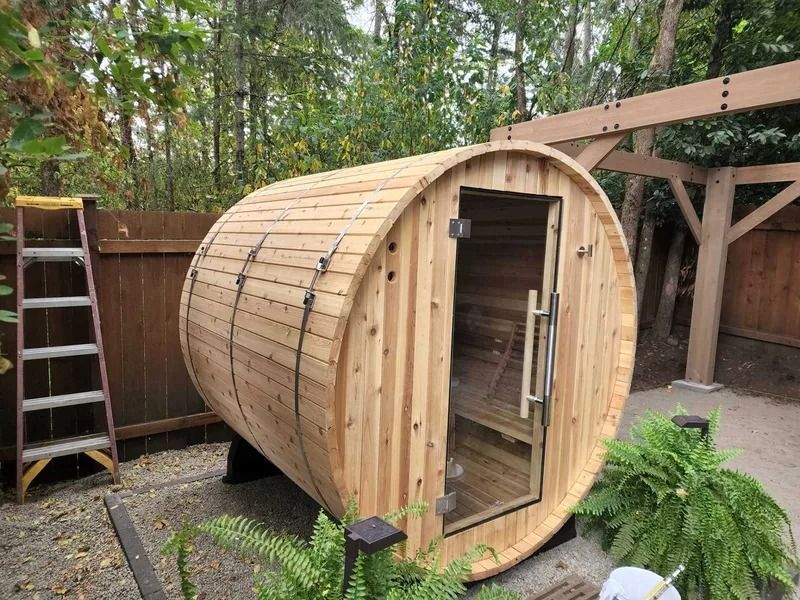 Wooden barrel sauna with glass door, outdoors near foliage, brown fence, and a ladder.