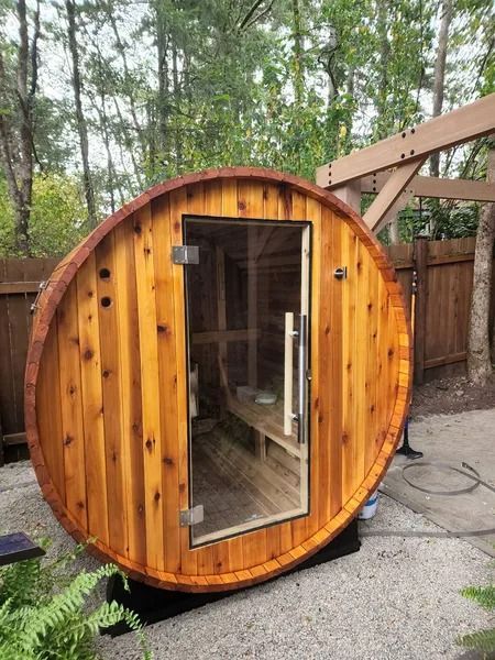 Wooden barrel sauna with glass door, set on a patio, with trees in the background.