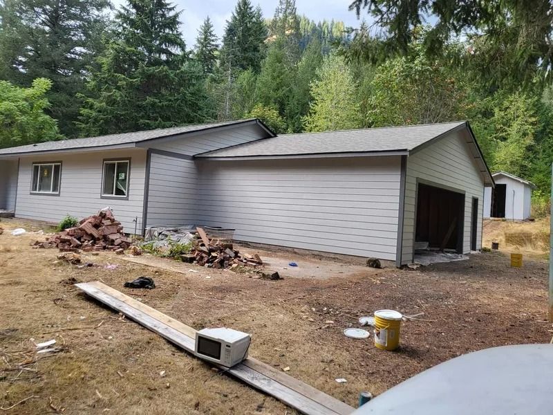 A house with attached garage under construction, with materials scattered in the yard and woods in the background.
