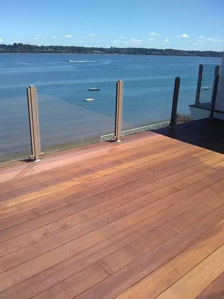 Wooden deck overlooking a calm blue body of water, glass railing, posts, distant boats, blue sky.