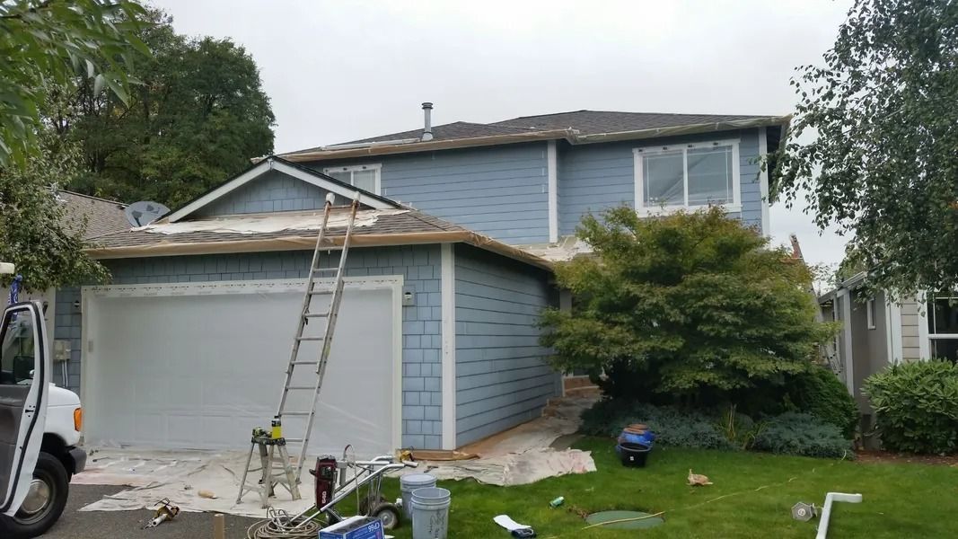 House exterior being painted, with ladder, garage door, blue siding, overcast sky, lawn.