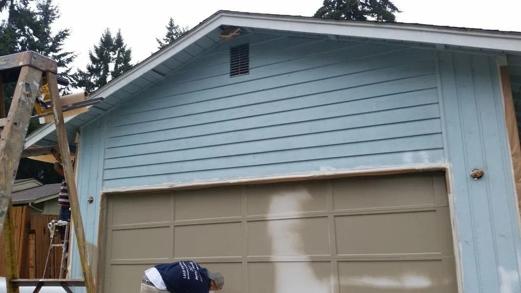 Man painting a tan garage door on a blue-sided building with a ladder in the yard.