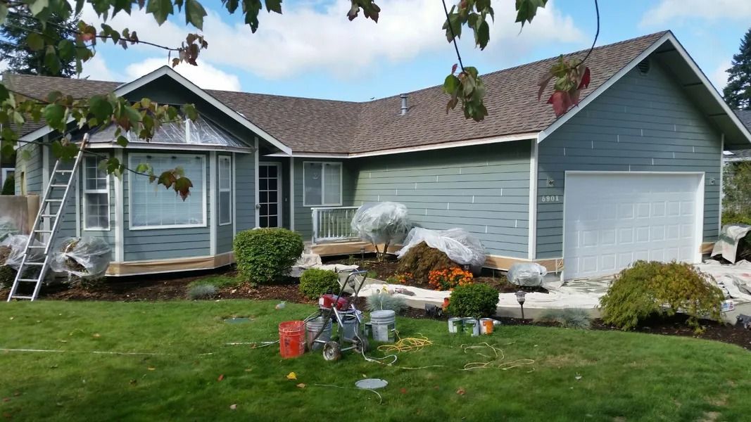 House being painted blue; ladder, paint supplies, and landscaping in front.