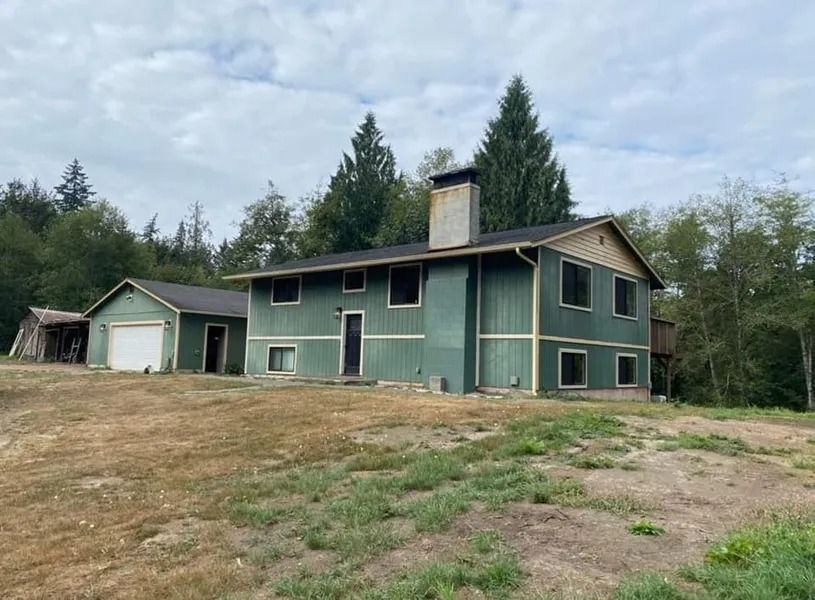 Green two-story house with a garage on a grassy hill, surrounded by trees under a cloudy sky.