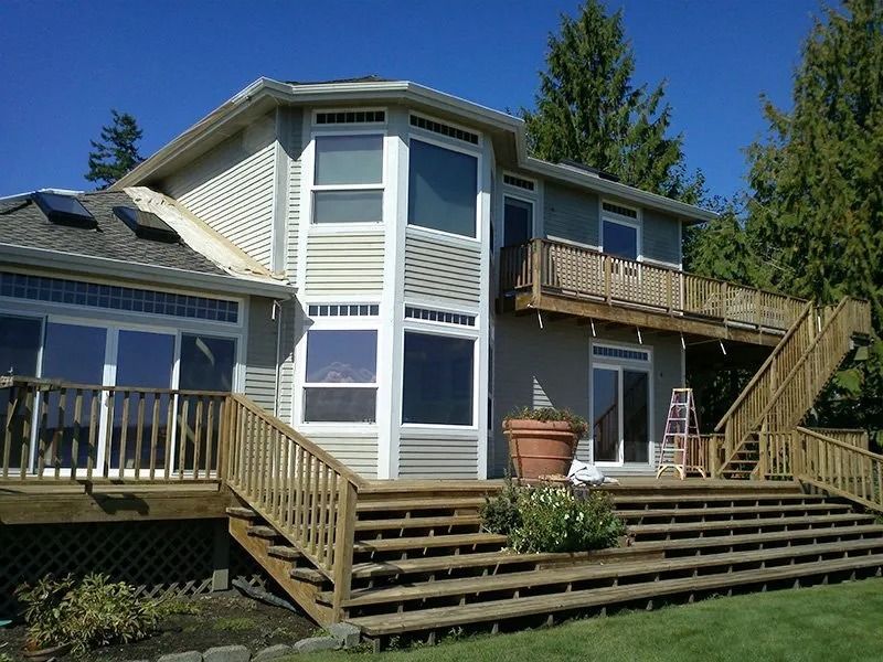 Two-story house with multi-level wooden decks and stairs, light siding, large windows, and a sunny outdoor setting.