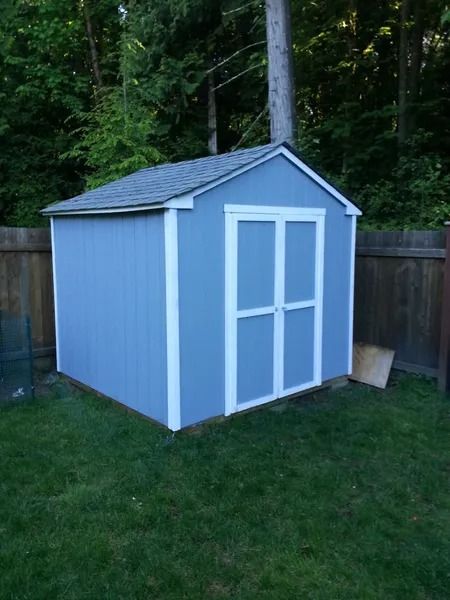 Blue shed with white trim, standing on grass, in front of a wooden fence and trees.