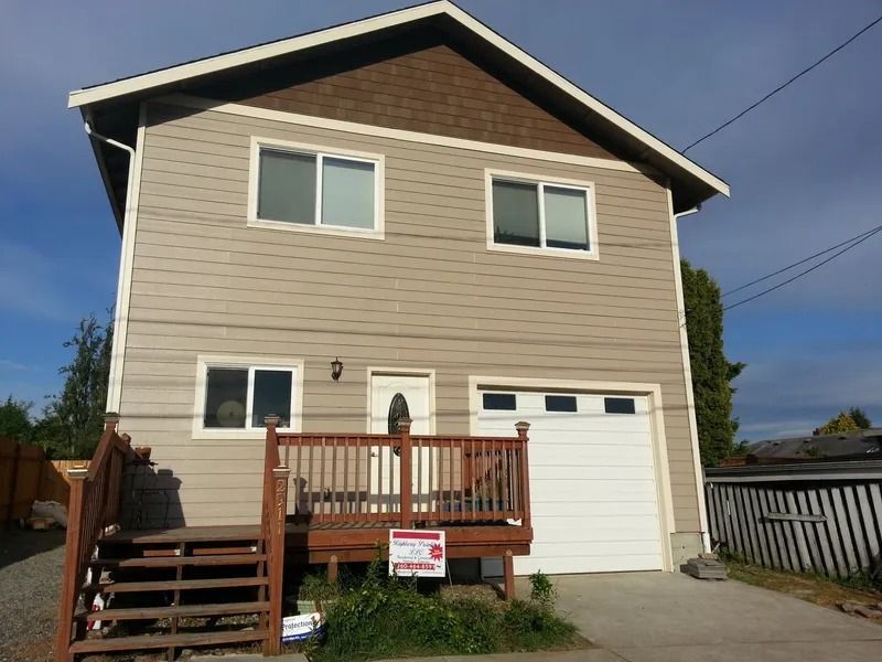 Two-story beige house with a brown roof and a white garage door, featuring a wooden deck and driveway.