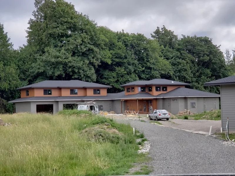 Circular, modern house under construction. Brown and gray exterior, dark roof, gravel driveway, and surrounding green grass.
