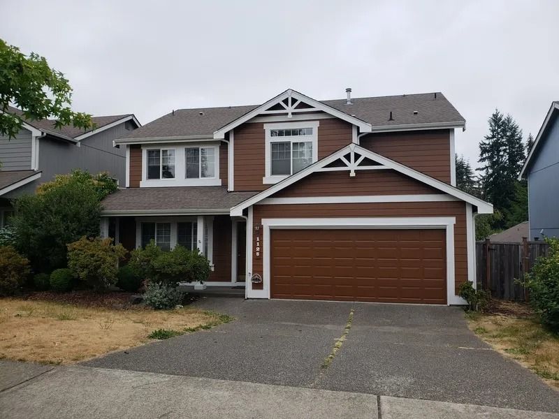 Brown two-story house with white trim, garage door, and driveway on a cloudy day.