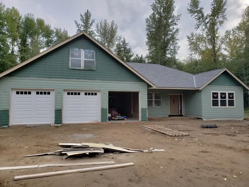 New house under construction, light green siding with white garage doors and a dark green roof.