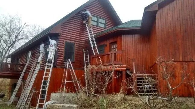 Workers painting a red log cabin exterior, using multiple ladders.