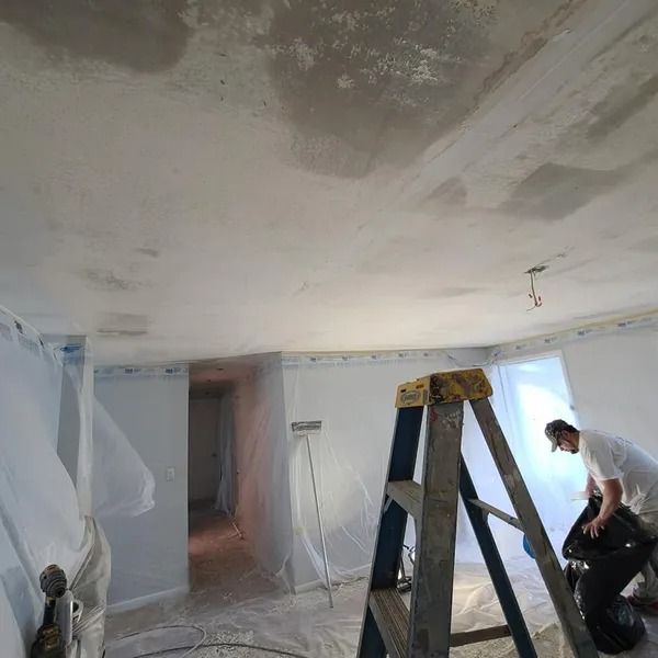 Room being renovated; ceiling with patches of concrete. Worker on ladder, others cleaning. Protective sheeting on walls.