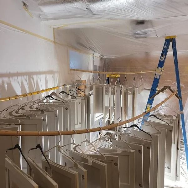 A room set up for painting, with white cabinet doors hanging on hangers from a wooden rod. A blue ladder is in the corner.