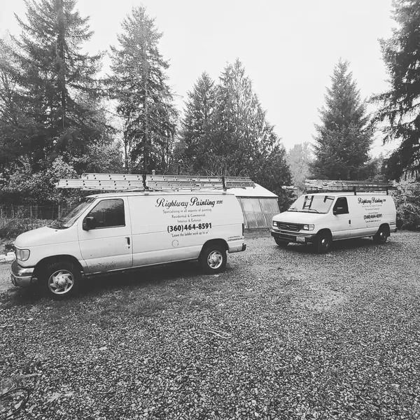 Two white work vans with ladders parked on gravel, trees in the background. 
