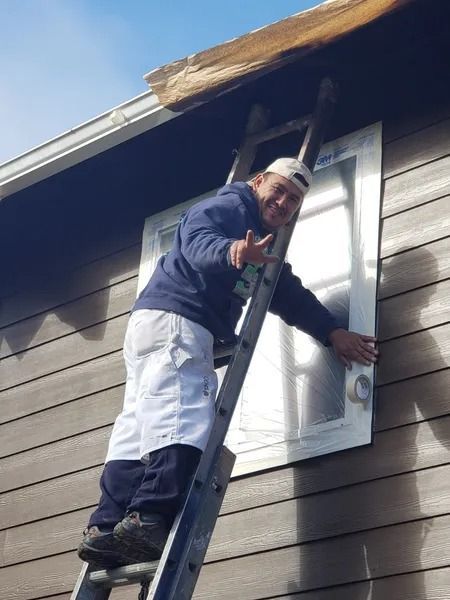 Man on ladder installing a window, smiling with a thumbs-up. Blue hoodie, white shorts, against gray siding.