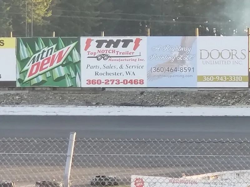 Billboard along a racetrack featuring advertisements for Mountain Dew, TNT Trucking, and Doors Unlimited.