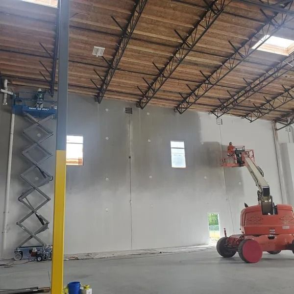 Construction workers on lifts install drywall inside a building with skylights and exposed beams.