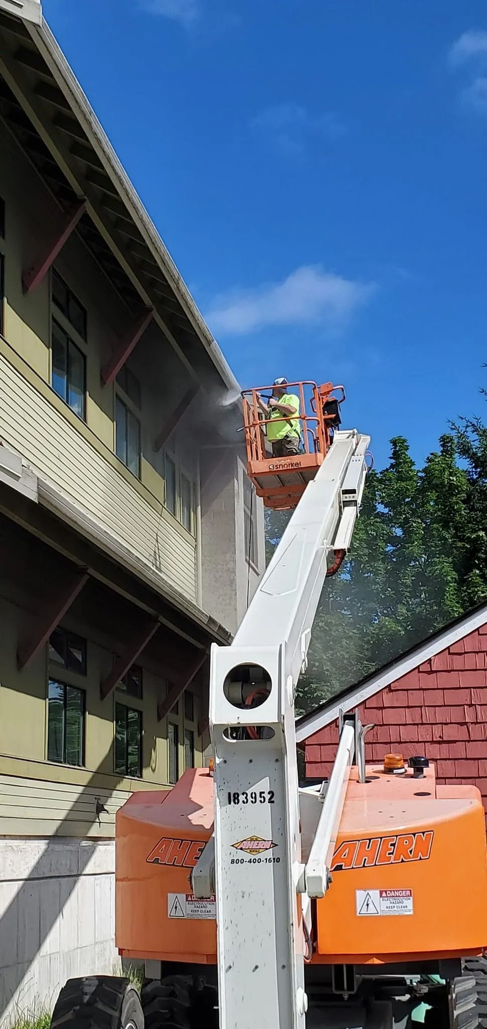 A worker in a lift spraying a house with water; a sunny blue sky is in the background.