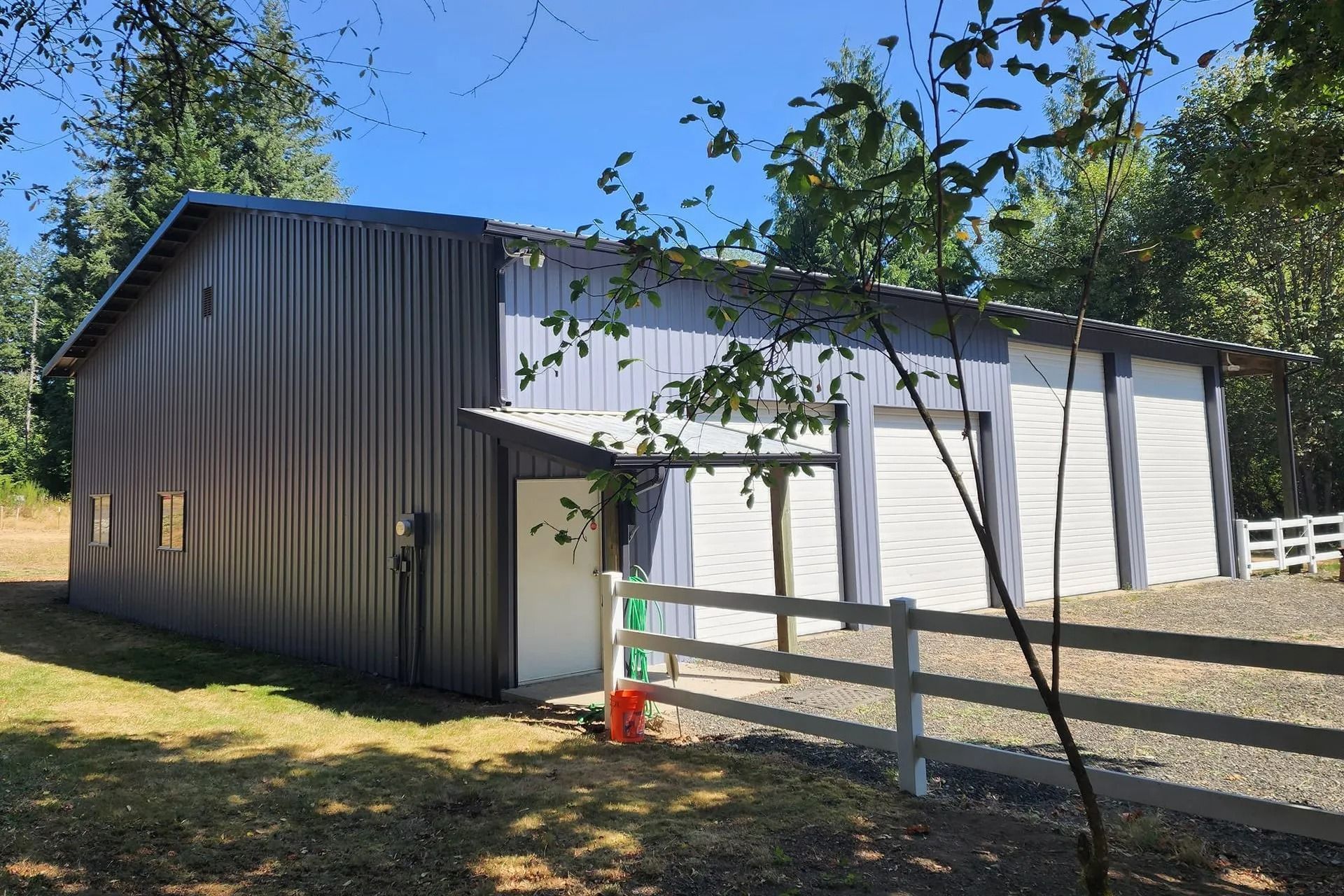 Gray and white metal barn with a driveway and white fence in a wooded area.