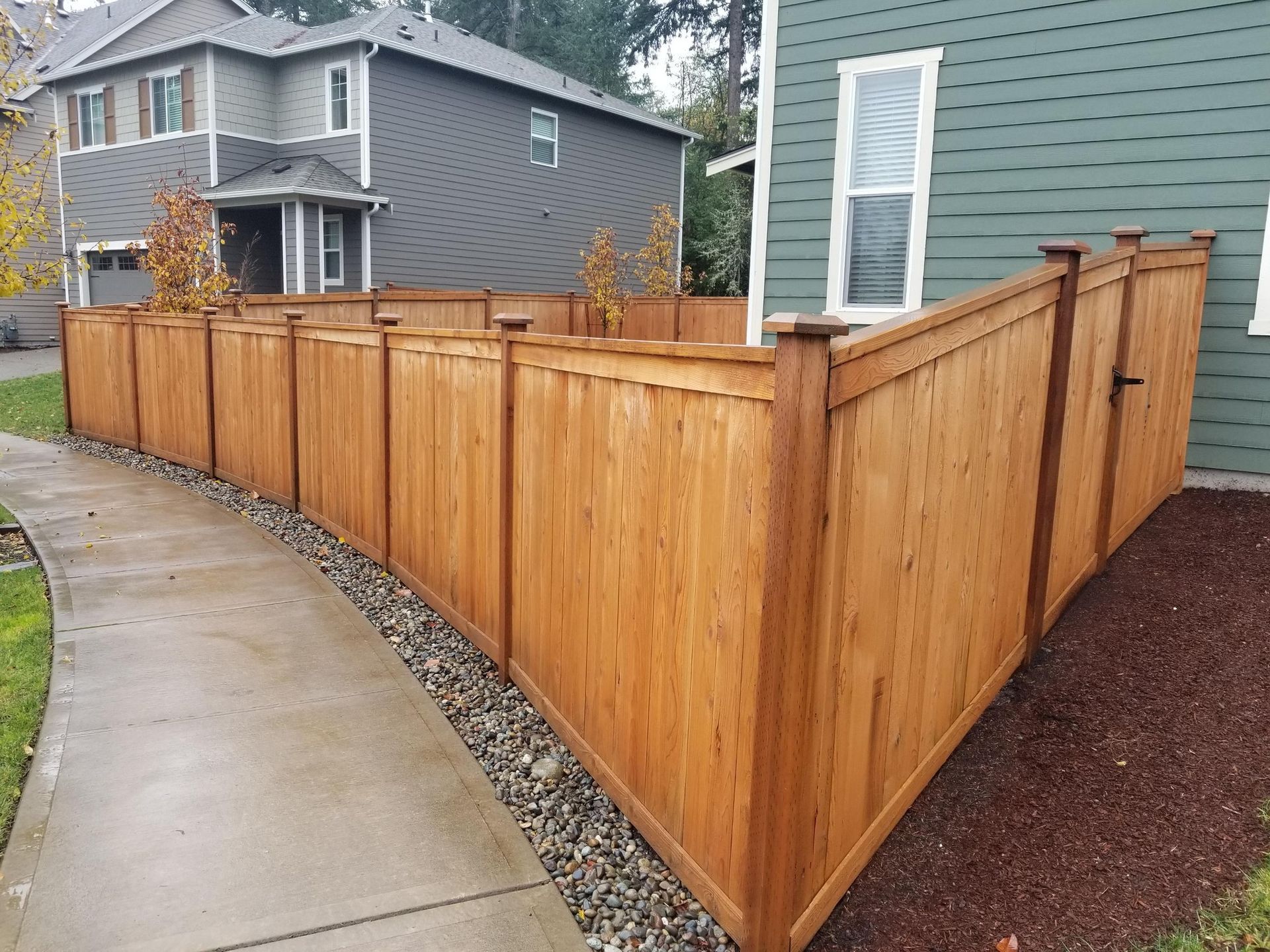 Wooden fence curves along a sidewalk, next to a teal house and grey house.