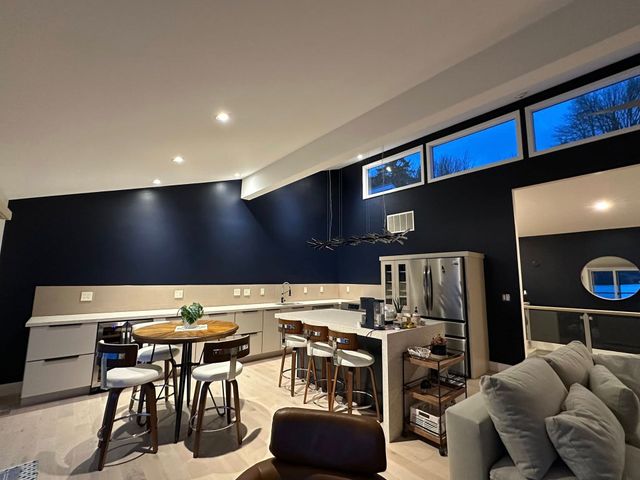 Modern kitchen with dark blue wall, island with stools, round table, and windows.