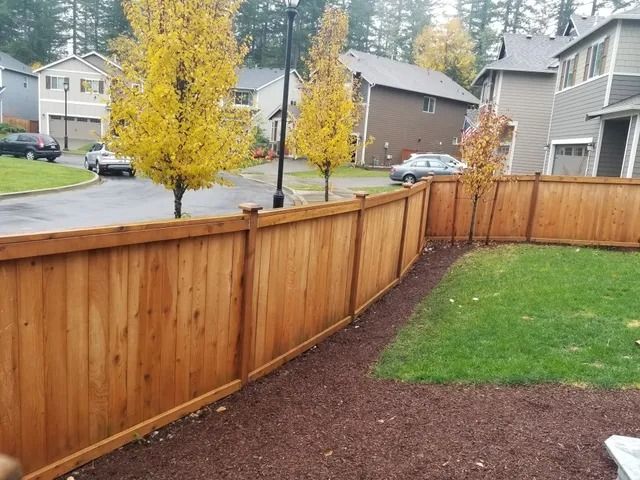 Wooden fence enclosing a backyard with green grass, brown mulch, and fall foliage.