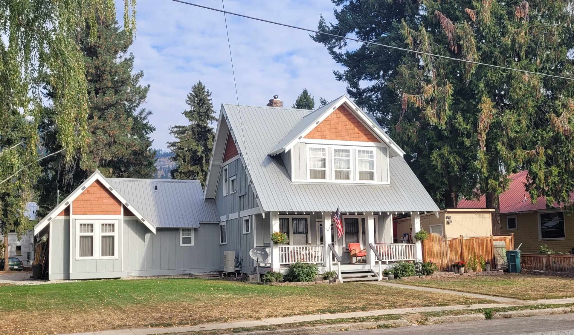 Gray house with brown trim and metal roof, small porch, lawn, trees, and blue sky.