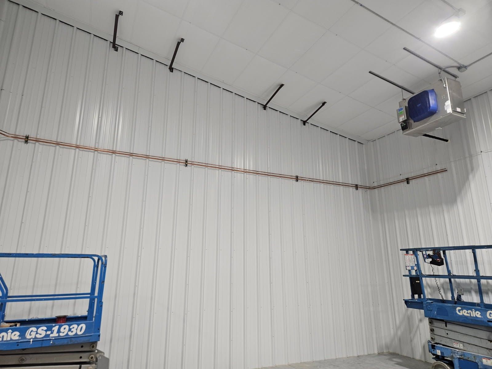 Interior view of a building with white walls, copper wiring, and an air handler mounted to the ceiling. Two blue lifts are visible.