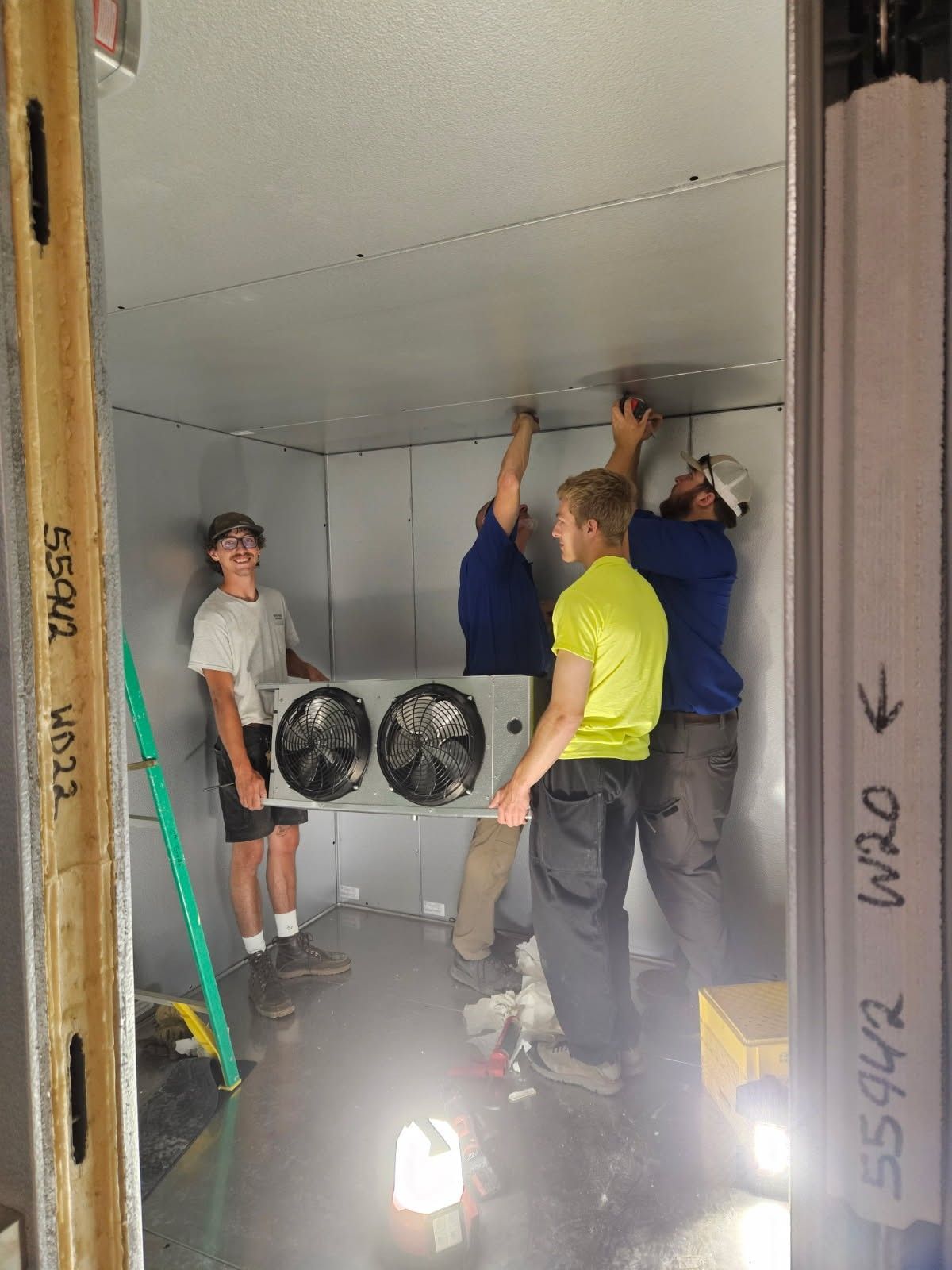 Four people installing a cooling unit inside a gray room, one person holds it up, others assist.