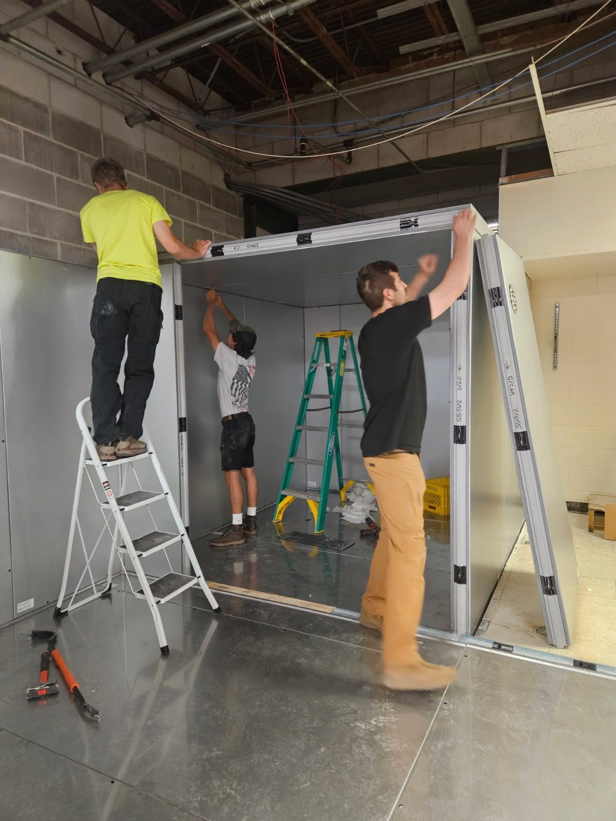 Three people constructing a metal enclosure inside a room. Two on ladders, one holding a large panel.