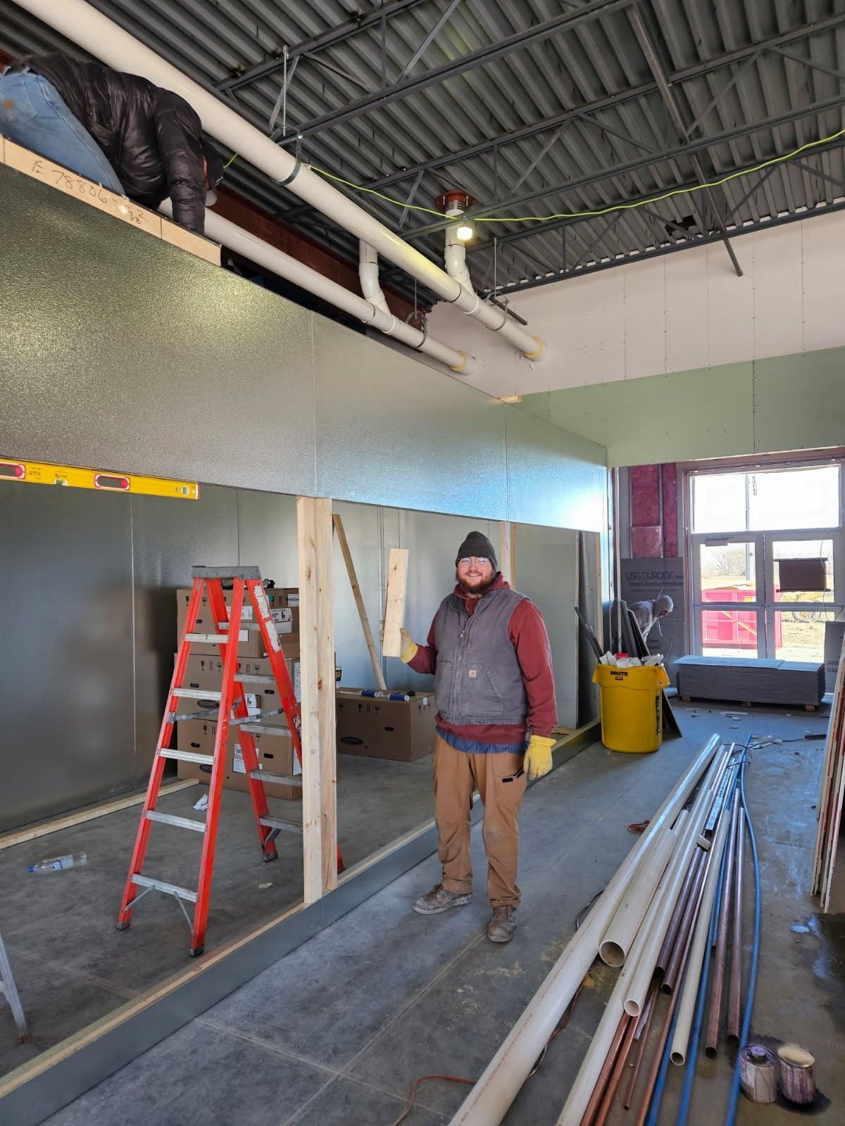 Construction worker in a building under renovation, holding wood, standing near a ladder. Piping and other supplies present.