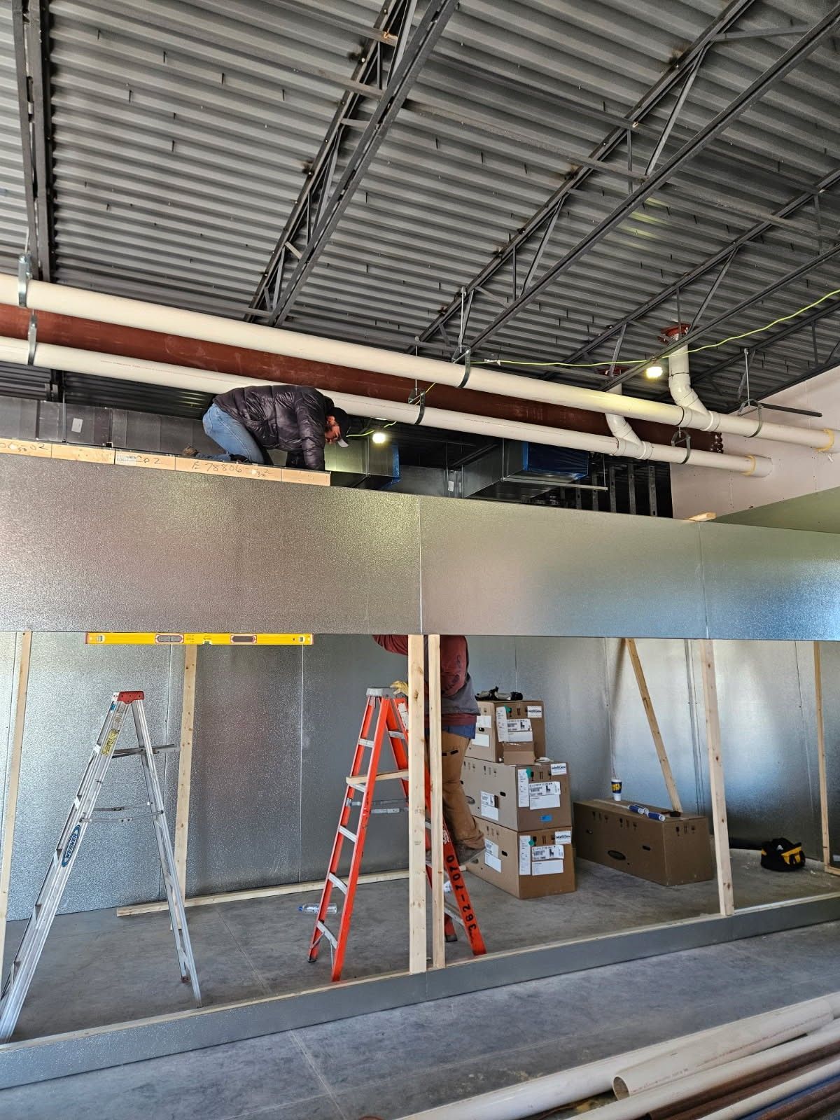 Construction site interior: worker on scaffolding installing pipes. Walls are framed and insulated.