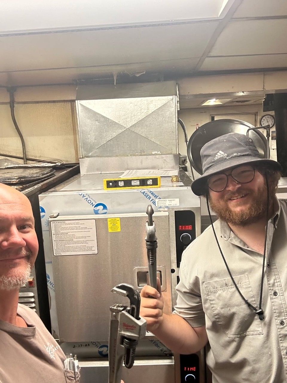 Two men in a kitchen; one holds a tool, smiling. A commercial oven is in the background.
