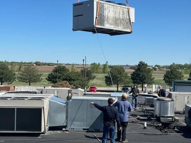 A rooftop HVAC unit being lifted by a crane; workers on roof point and supervise on a sunny day.