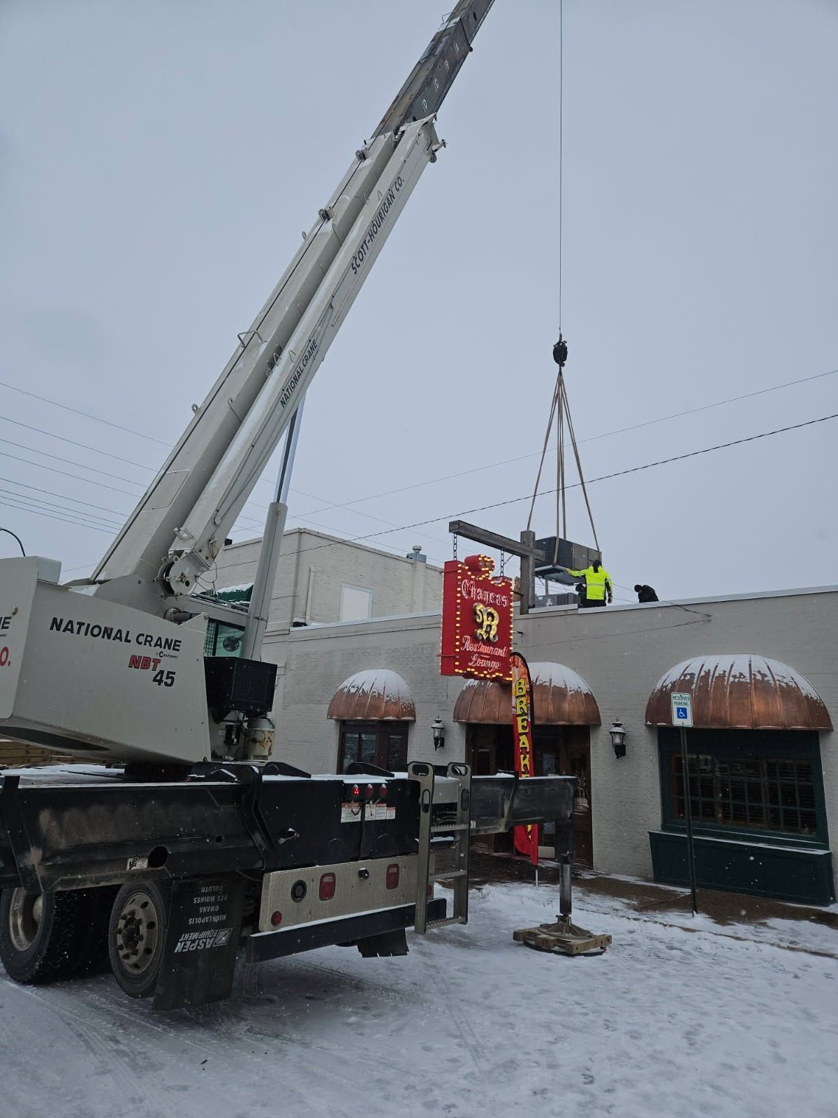 Crane lifting a sign onto a snowy building. A person in a high-vis vest stands on the roof.