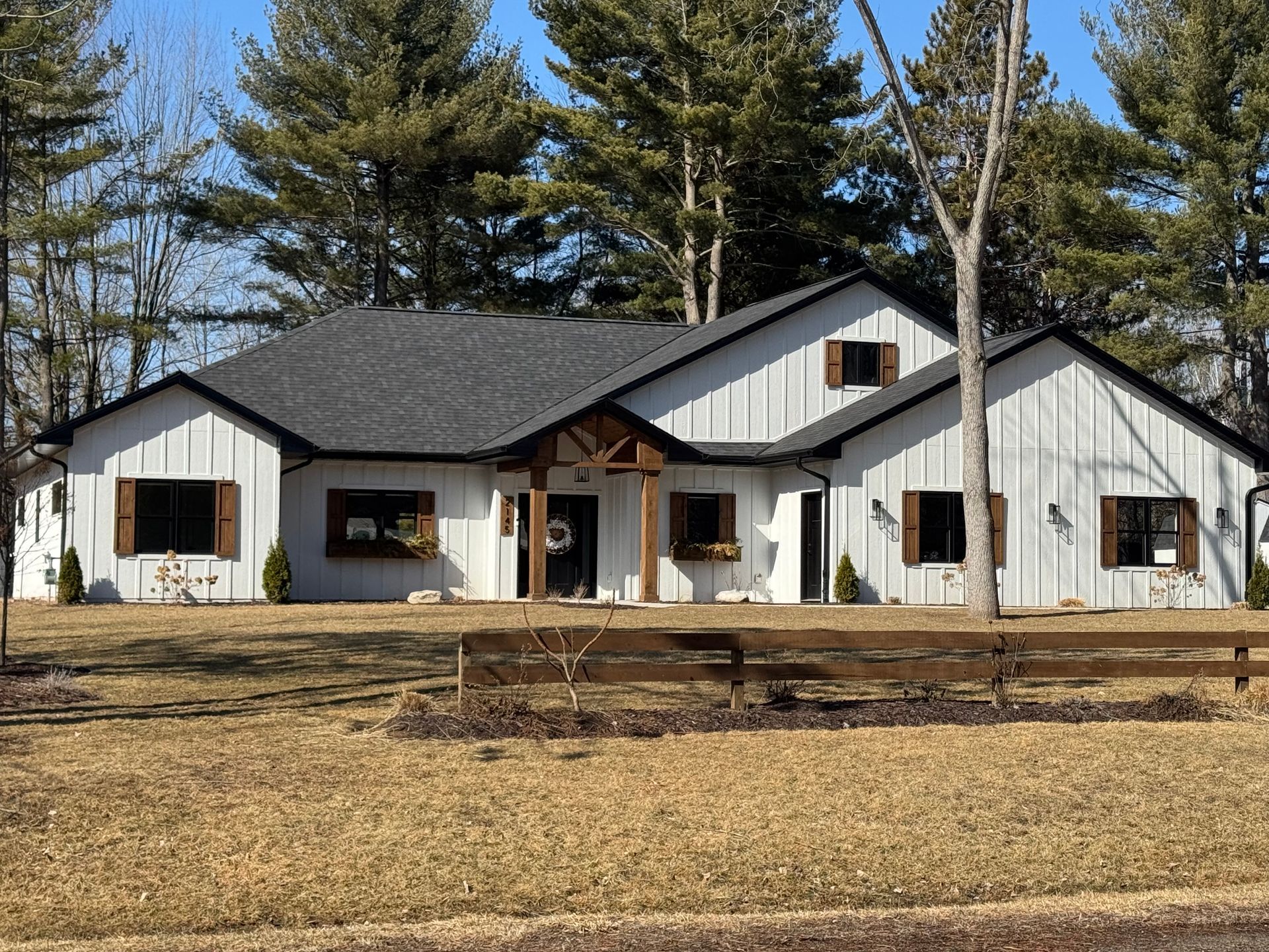 A large white house with a black roof is surrounded by trees and a wooden fence.