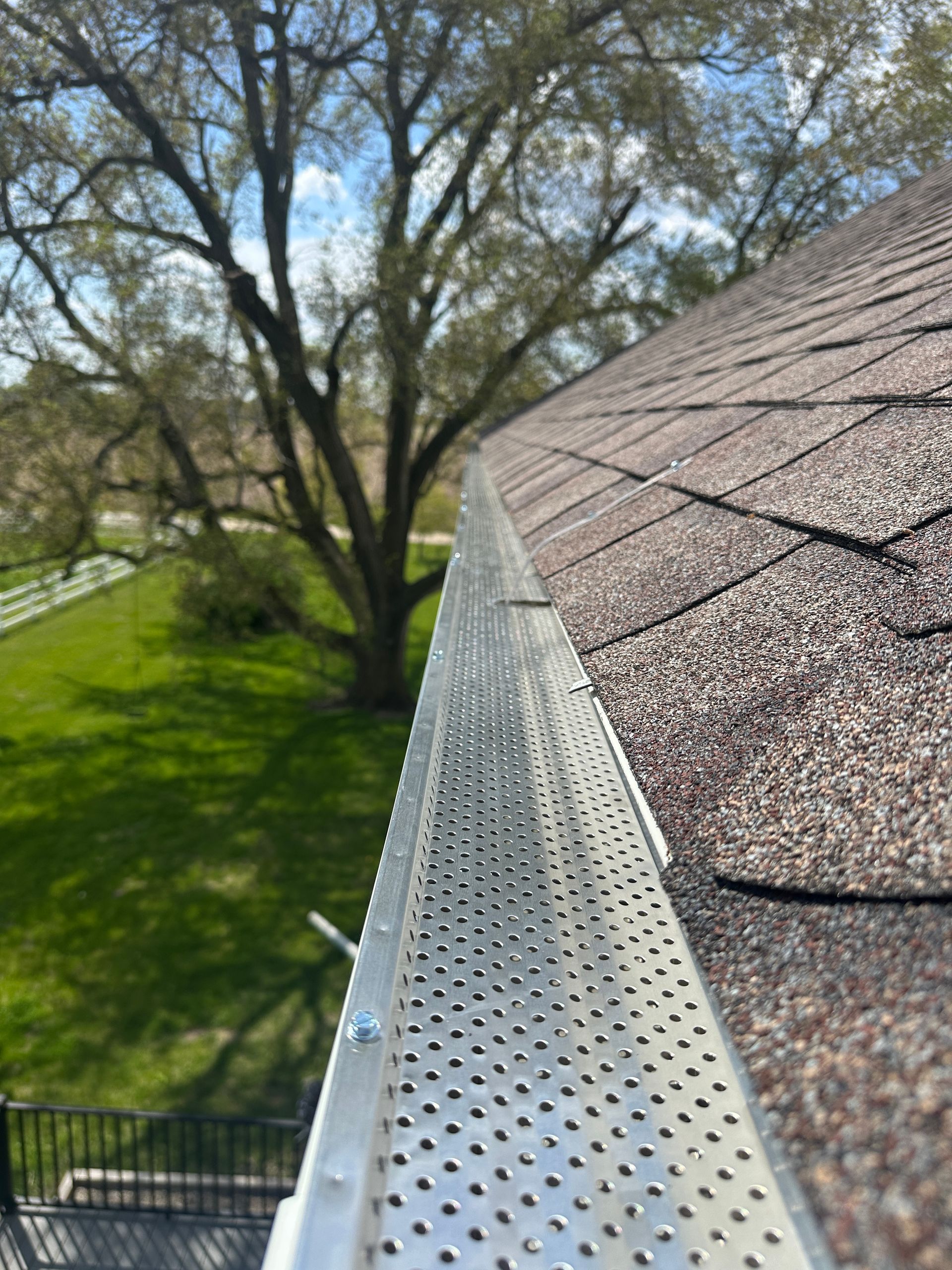 A metal perforated gutter guard installed on a residential roof edge, with a tree and grassy yard in the background.