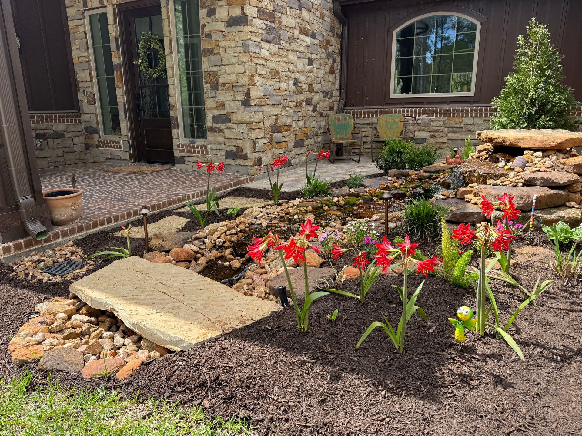 Flower bed with red blooms and stone edging beside a brick house and walkway