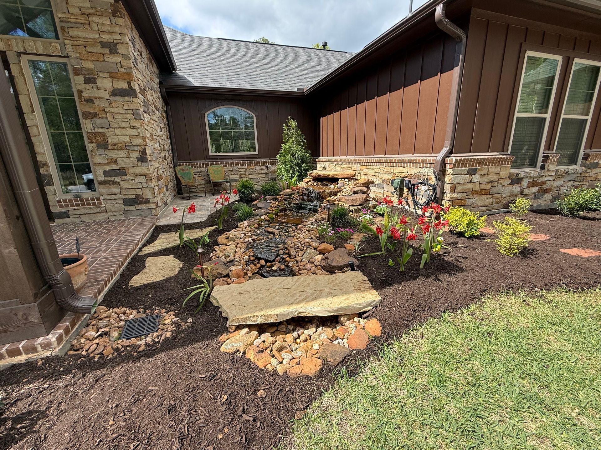 Stone house front yard with a curved flower bed, mulch, rocks, and blooming red and white flowers